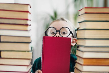 Büyük gözlüklü anaokulu kızı kırmızı kitapların arasında fotokopi alanı olan kırmızı kitaplara bakıyor.