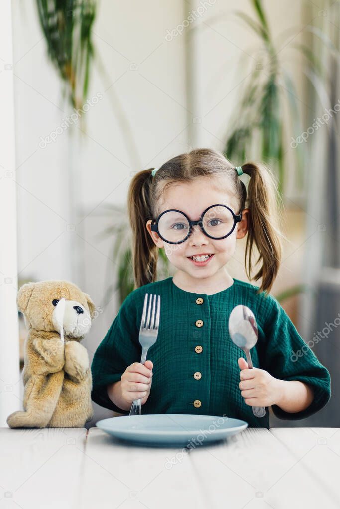 Little beautiful girl in green muslin dress waiting breakfast with bear toy. Little cute girl at white dining table in kitchen. Healthy nutrition for young kids. Good morning concept