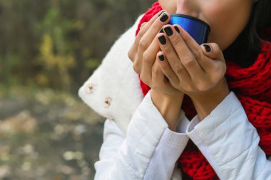 Beyaz önlüklü, tığ işi kırmızı atkı ve siyah şapkalı bir kadın elinde bir bardak tutuyor. Doğada çay partisi