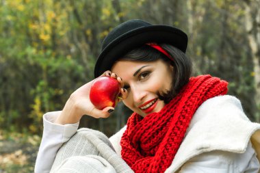 Siyah şapkalı, kırmızı eşarplı, elinde kırmızı elmalı bir kadın gülümsüyor ve kameraya bakıyor..