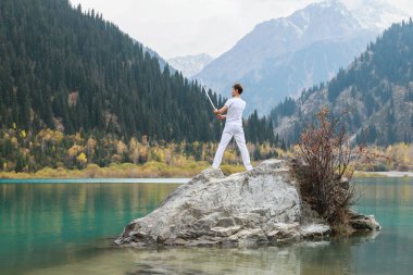Genç bir adam büyük bir taşın üzerinde duruyor ve elinde bir Japon kılıcı tutuyor..