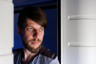 Sakallı yakışıklı, beyaz bir adam, trenin penceresinden dışarı bakıyor, sıkılmış ya da yorgun görünüyor.