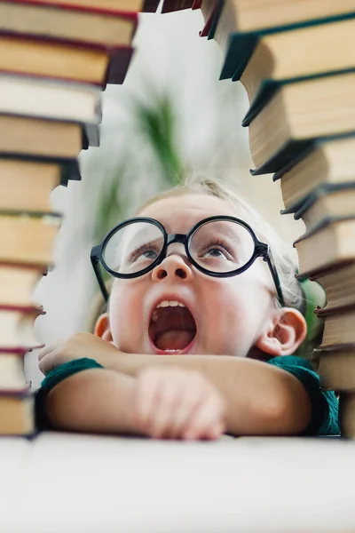 Shocked little cute blonde kid girl 4-6 years old in library among many books. Home education concept. Vertical — Stock Photo, Image