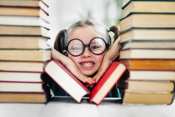 Preschool age girl with wondering face in big funny glasses among a pile of books.