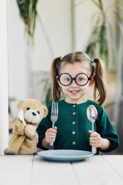 Oyuncak ayıyla kahvaltı bekleyen tatlı bir kız. Mutfaktaki beyaz yemek masasındaki küçük tatlı kız. Genç çocuklar için sağlıklı beslenme.