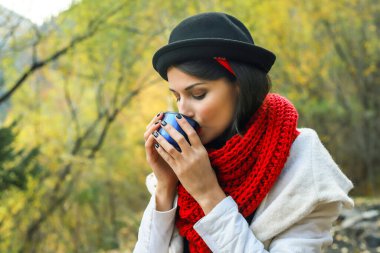 Beyaz önlüklü, tığ işi kırmızı atkı ve siyah şapkalı bir kadın elinde bir bardak tutuyor. Doğada çay partisi