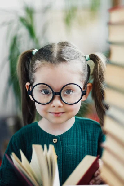 Cute preschool age girl with dimples on her cheeks in round big glasses looking at a camera between two books piles — Stock Photo, Image