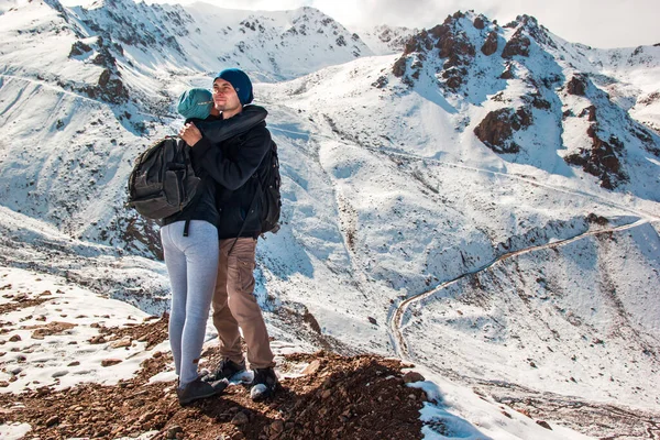 Um jovem abraça sua esposa em um fundo de montanhas nevadas. Férias nas montanhas . — Fotografia de Stock