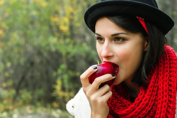 Beautiful woman eating a ripe red apple. Crochet red scarf and black hat, outdoor picnic