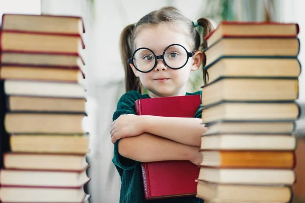 Fille d'âge préscolaire dans de grands verres tenant le livre rouge parmi les grappes de livres — Photo