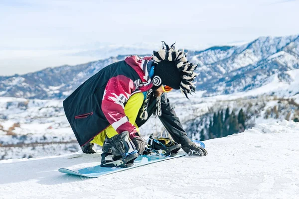 O snowboarder de homem jovem no chapéu fanny senta-se na neve e fixa fixações em um snowboard antes da descida — Fotografia de Stock