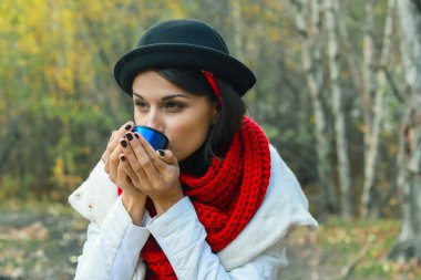 Beyaz önlüklü, tığ işi kırmızı atkı ve siyah şapkalı bir kadın elinde bir bardak tutuyor. Doğada çay partisi