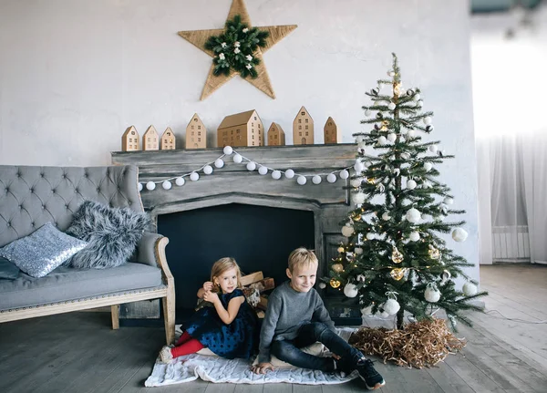 children - a boy and a girl are sitting by the fireplace, a New Year tree, playing with cardboard houses, a cozy Christmas decor and children\'s emotions