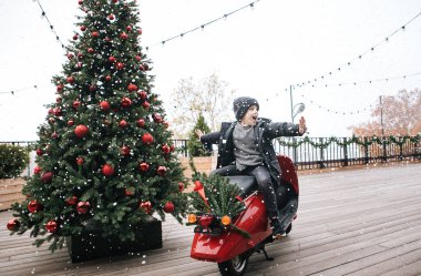 kış giysileri içinde bir oğlan yakınındaki yeni yıl ağacı ahşap bir teras üzerinde kırmızı bir motosiklet üzerinde oturuyor, bu kar yağıyor, neşeli çocuk duygular, Noel için fotoğraflar