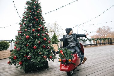 kış giysileri içinde bir oğlan yakınındaki yeni yıl ağacı ahşap bir teras üzerinde kırmızı bir motosiklet üzerinde oturuyor, bu kar yağıyor, neşeli çocuk duygular, Noel için fotoğraflar