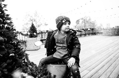 a boy in winter clothes is sitting on a red motorcycle near the New Year tree on a wooden terrace, it is snowing, joyful children's emotions, photos for Christmas clipart