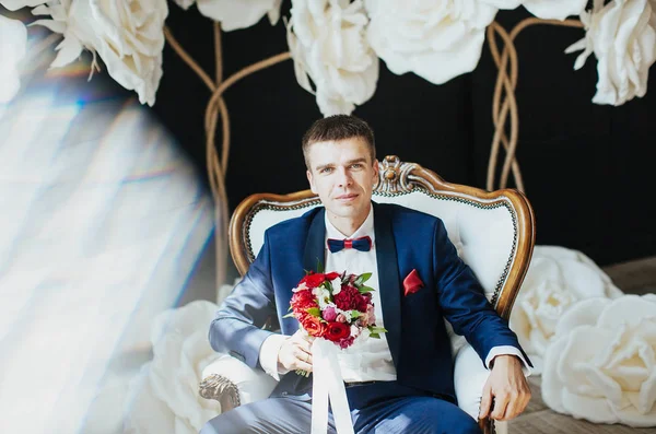 the groom in a stylish blue suit is sitting on a white vintage chair - a wedding photo in the studio on a background of white huge flowers