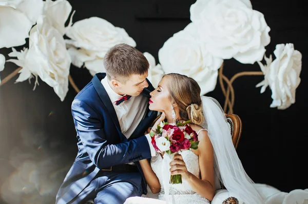 穿着漂亮礼服的新娘和新郎坐在白色的老式椅子上拥抱 在演播室里 在巨大的白花背景下合影 — 图库照片
