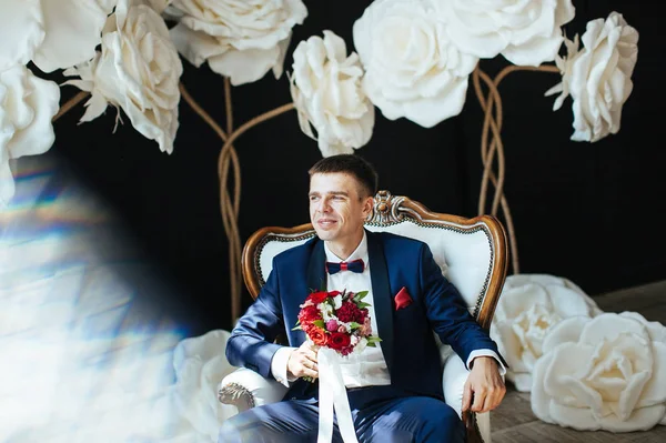 the groom in a stylish blue suit is sitting on a white vintage chair - a wedding photo in the studio on a background of white huge flowers