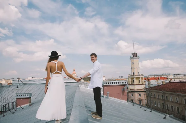 loving couple - a girl in a white dress and a hat and a guy - on a walk on the roof hugging and laughing, a view from the height of the city, positive and sincere emotions of love