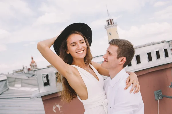 有爱心的一对 一个穿着白色衣服 帽子和男人的女孩 在屋顶上散步 拥抱和笑 从城市的屋顶上看到的是积极而真诚的爱的情感 — 图库照片