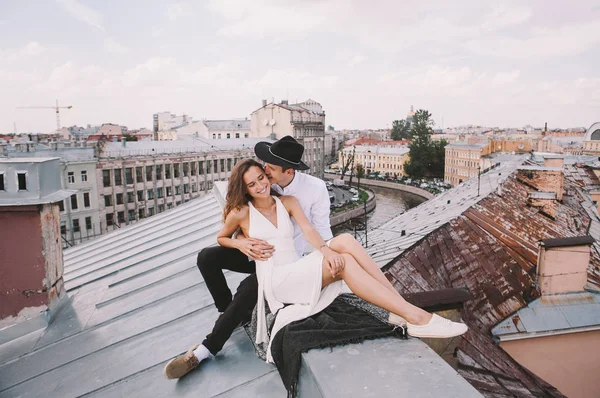 loving couple - a girl in a white dress and a guy with a hat - on a walk on the roof hugging and laughing, a view from the roof of the city, positive and sincere emotions of love