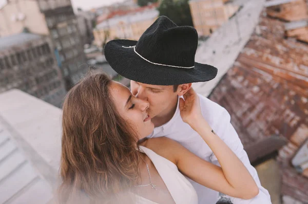 有爱心的情侣 一个穿着白色衣服的女孩和一个戴帽子的男人 在屋顶上散步 拥抱和笑 从城市的屋顶上看到的是积极而真诚的爱的情感 — 图库照片