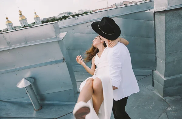 loving couple - a girl in a white dress and a guy with a hat - on a walk on the roof hugging and laughing, positive and sincere emotions of love