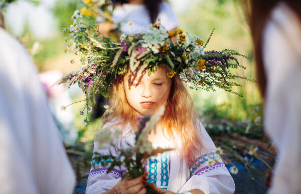 cute girl blonde in white national dress weaves a wreath of flowers and grass at the pagan festival of spring