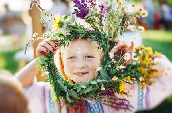 cute girl blonde in white national dress weaves a wreath of flowers and grass at the pagan festival of spring