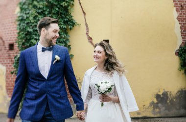 sarı duvar ve sarmaşık arka planına karşı bir yaz parkında yürürken eller tutarak ve gülümseyen onların düğün gününde şık gelin ve damat