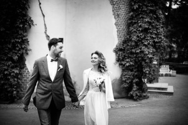 duvar ve sarmaşık, siyah beyaz fotoğraf arka planına karşı bir yaz parkında yürürken eller tutarak ve gülümseyen onların düğün gününde şık gelin ve damat 