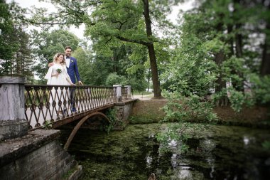 bir taş köprüde bir yazlık parkta yürürken kucaklayan ve gülümseyen düğün gününde şık gelin ve damat