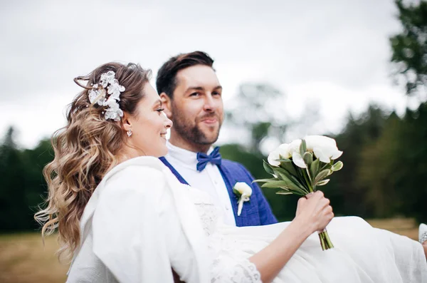 フィールドで腕に彼の幸せな花嫁を運ぶ新郎 珍しい結婚式の日 — ストック写真