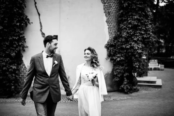 时尚的新郎新娘在婚礼当天手牵手 微笑着走在一个夏天的公园里 背景是墙和常春藤 黑白照片 — 图库照片