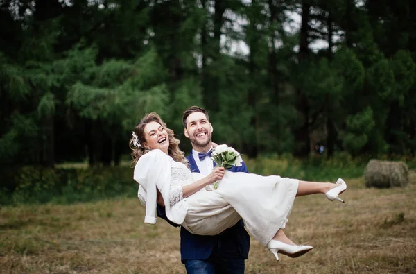 フィールドで腕に彼の幸せな花嫁を運ぶ新郎 珍しい結婚式の日 — ストック写真