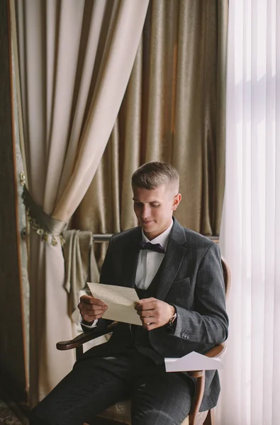 早晨时尚新郎 一个年轻的家伙金发在灰色夹克读一封信从新娘在复古房间 — 图库照片
