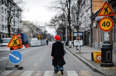 Kırmızı şapkalı, ceketli ve atkılı renkli sarışın kız şehirde yürürken gülümsüyor.