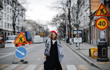 Kırmızı şapkalı, ceketli ve atkılı renkli sarışın kız şehirde yürürken gülümsüyor.