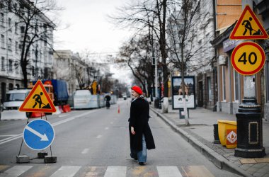 Kırmızı şapkalı, ceketli ve atkılı renkli sarışın kız şehirde yürürken gülümsüyor.