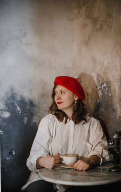 Beyaz tişörtlü, kırmızı bereli, eski moda bir iç mekânda poz veren ve gülümseyen güzel sarışın kız.