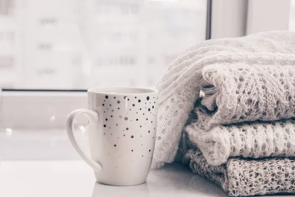 Stack of cozy knitwears and a cup of coffee on white marble windowsill against white window background. Copy space.