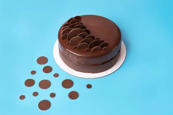 Dark chocolate mousse cake covered with mirror glaze and decorated with round chocolate chips on blue background. Modern stylish cake. Copy space.