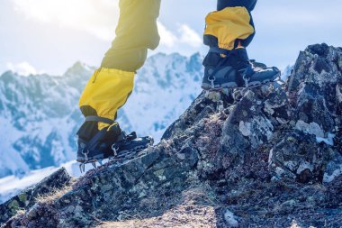 Climber in crampons stands on the rocks in front of the entrance to the peak on the background of the snowy mountains. Feet close up. The concept of the travel path and achieving the goal clipart