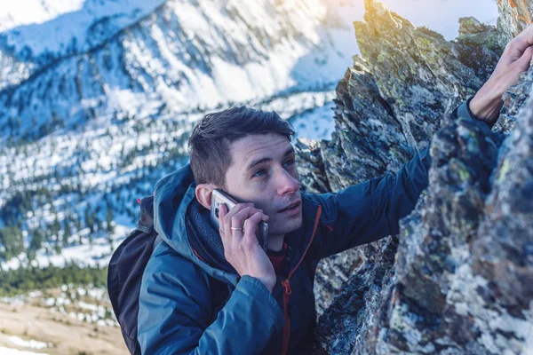 A young man climbs the mountain to the top and talking holding the phone in his hand. The concept of persistence and goal achievement in your career. Work in difficult conditions