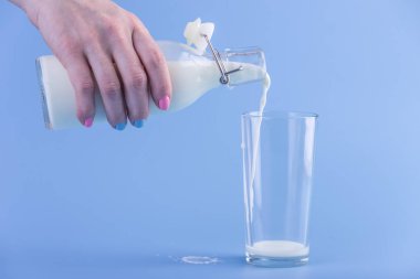 Bir kadın eli mavi bir arka plan üzerine bir bardak içine bir cam şişe taze süt dökülen. Renkli minimalizm. Kalsiyum sağlıklı süt ürünleri kavramı