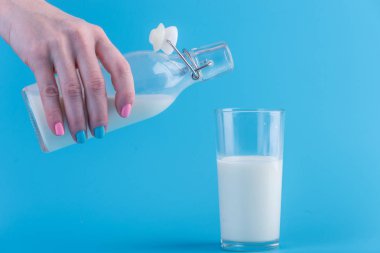 Bir kadın eli mavi bir arka plan üzerine bir bardak içine bir cam şişe taze süt dökülen. Renkli minimalizm. Kalsiyum sağlıklı süt ürünleri kavramı