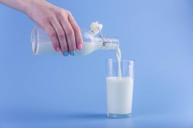 Bir kadın eli mavi bir arka plan üzerine bir bardak içine bir cam şişe taze süt dökülen. Renkli minimalizm. Kalsiyum sağlıklı süt ürünleri kavramı