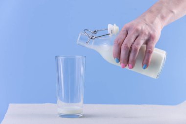 Bir kadın eli mavi bir arka plan üzerine bir bardak içine bir cam şişe taze süt dökülen. Renkli minimalizm. Kalsiyum sağlıklı süt ürünleri kavramı