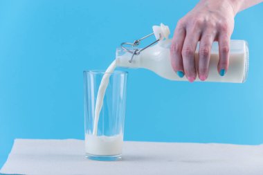 Bir kadın eli mavi bir arka plan üzerine bir bardak içine bir cam şişe taze süt dökülen. Renkli minimalizm. Kalsiyum sağlıklı süt ürünleri kavramı